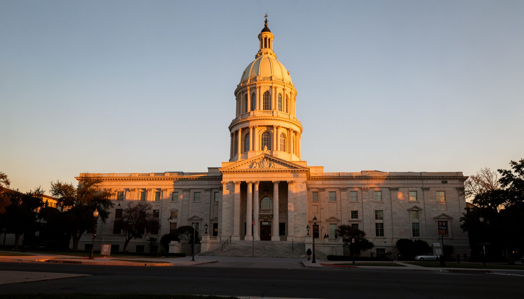 A wide photograph of the Texas State Capitol building in Austin at golden hour, warm amber light washing over the limestone façade against a soft blue sky.